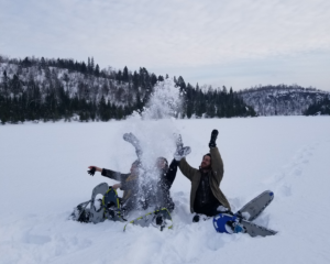 Three people sit in the snow and throw snow up into the air. There is a beautiful wintery forest landscape behind them, and snowshoes in the snow beside them.
