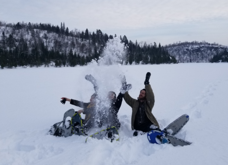 Three people sit in the snow and throw snow up into the air. There is a beautiful wintery forest landscape behind them, and snowshoes in the snow beside them.