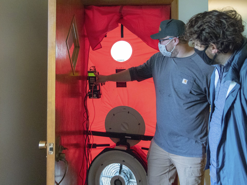 Two people look at a blower door setup: red fabric stretched inside a door frame, with a fan and monitoring system.