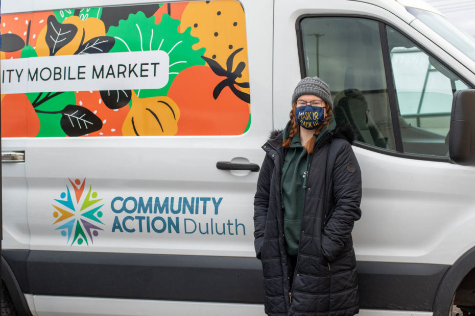 Person in winter gear and face mask stands in front of a van with "Community Action Duluth Mobile Market" on the side