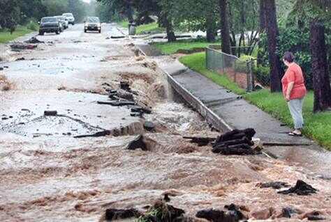 Person standing on a sidewalk looks down a road that is filled with rushing floodwater. Chunks of asphalt and pavement stick up out of the water, and the street appears crooked.