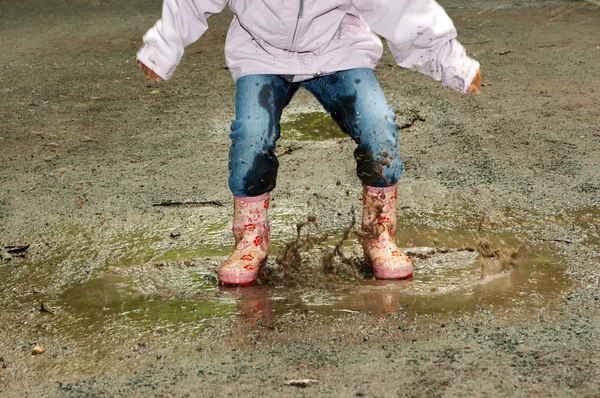 Child stomps in mud puddle wearing muddy yellow rainboots. We see shoulders down, but we can tell they are happy from their energetic pose.