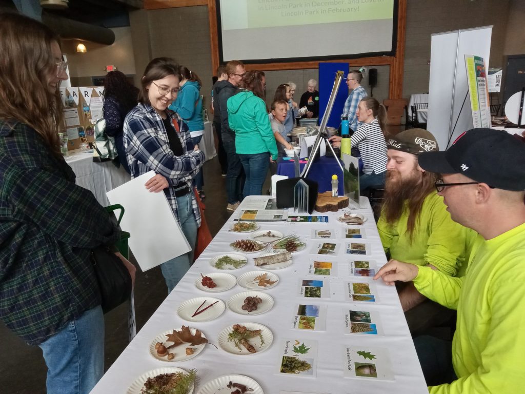 Two standing women smile and talk with two men seated at a table. The table has photos of local trees and small parts of leaves, branches, etc. for a matching activity.