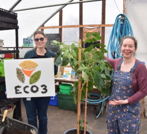 Two women pose in a greenhouse with a large plant and a sign depicting the Eco3 logo in seeds.
