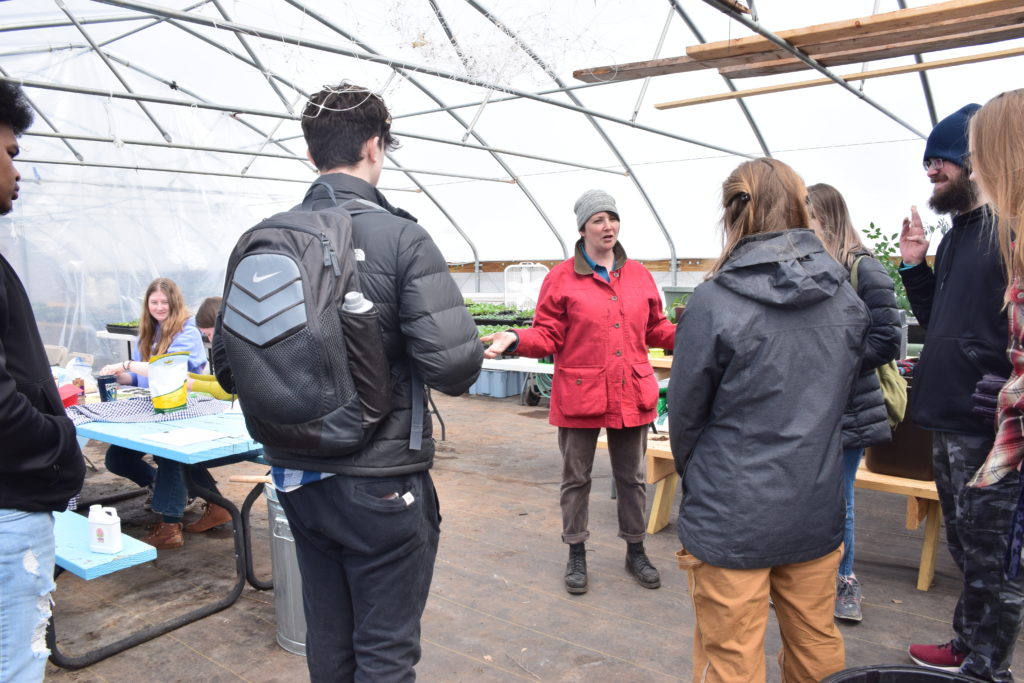 Woman in red coat and winter cap faces the camera, gesturing as she talks. She is addressing a small group of college students, some with backpacks, who are facing away from the camera. They are all in a large greenhouse, with a few plants in the background.