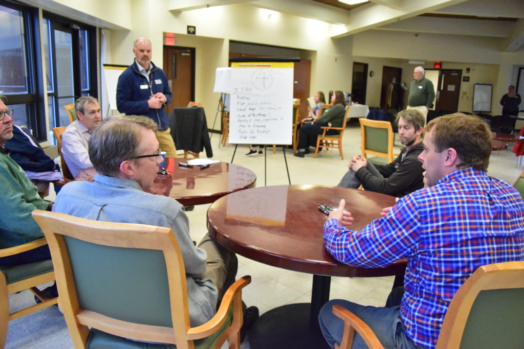 Two men sitting at a table talk with each other. In the background, a man is standing by a board where ideas are being recorded (text is too small to read).