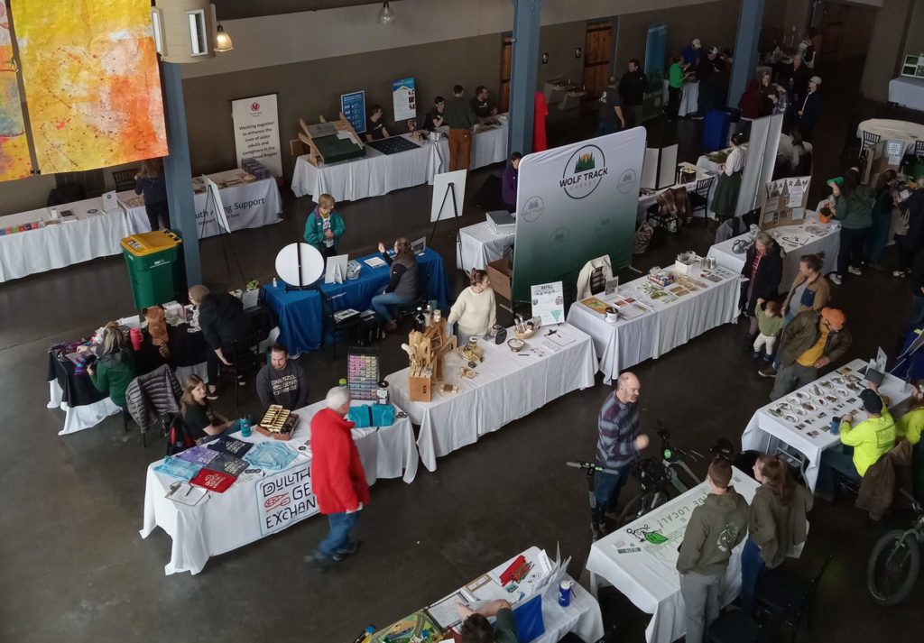 From a balcony above the action, we see many tables with displays and people talking and walking around at EcoFest.