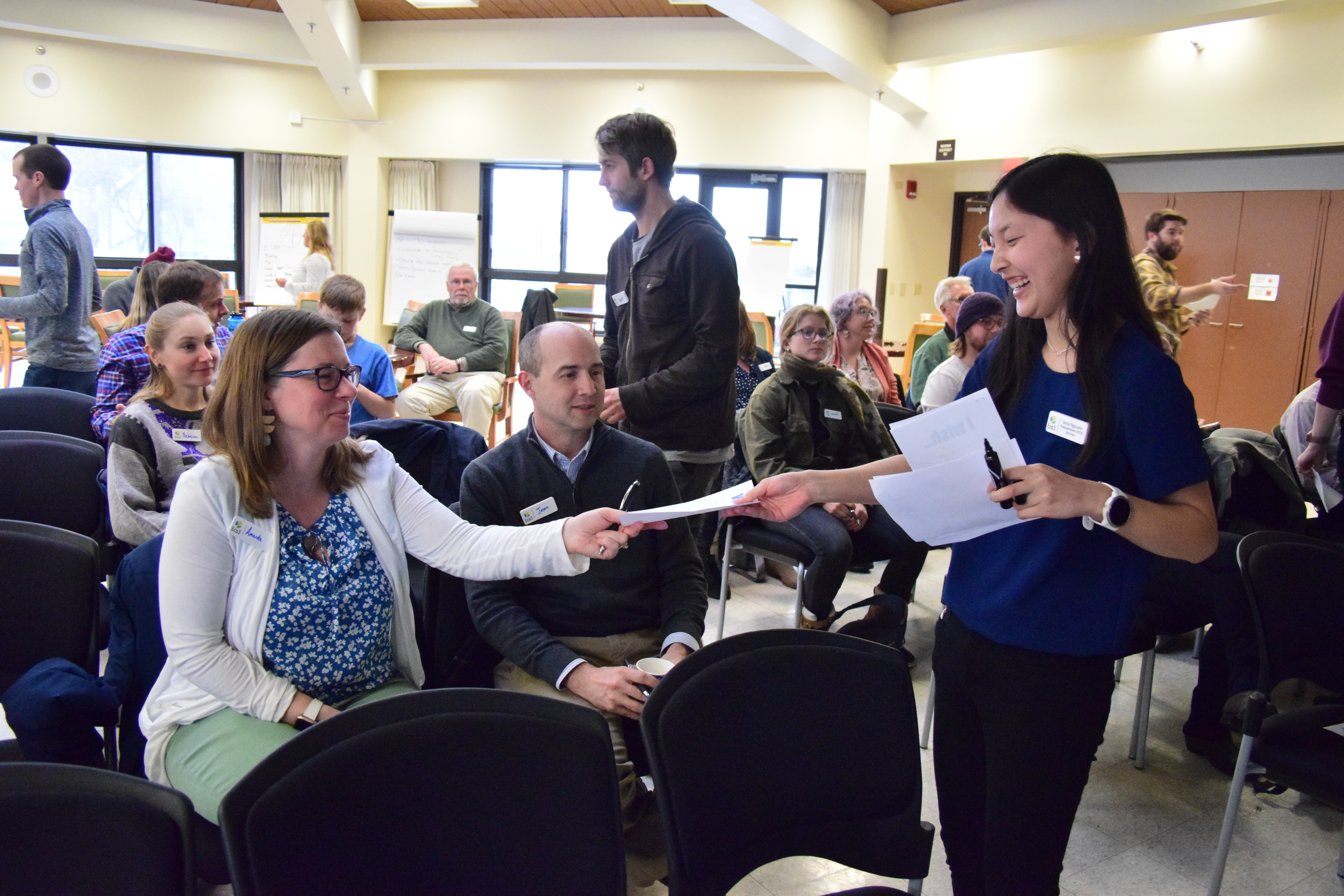 Several people are seated in chairs facing the camera. A few people walk among the chairs to gather papers from the seated audience. A young woman smiles as she takes a paper from a seated woman.