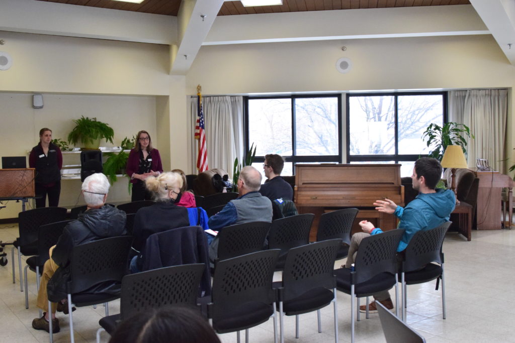 Two women stand at the front of a room. The are several people seated in chairs facing them. A man in a chair is gesturing as he speaks to the women.