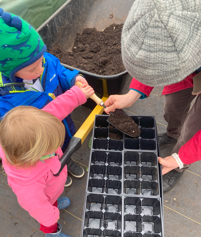 Woman helps two children in winter clothing put dirt into small containers for planting seeds.