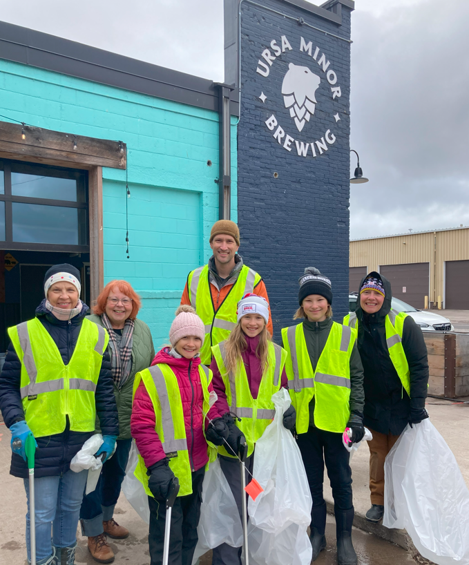 People of all ages wearing high-visibility vests stand in front of the Ursa Minor Brewing sign and smile. They are holding trash bags and other cleanup materials.