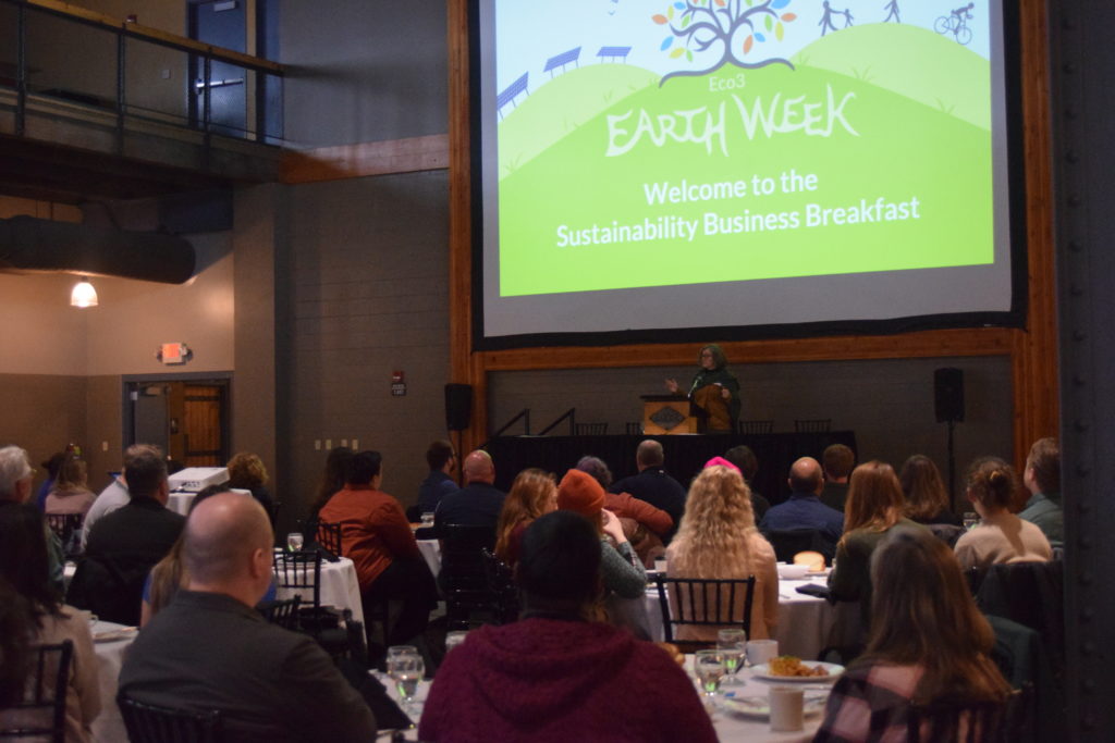 Michele Reeves stands at a podium and speaks to a crowd of seated people, who are facing away from the camera. The Earth Week logo is displayed on a screen on the back wall.