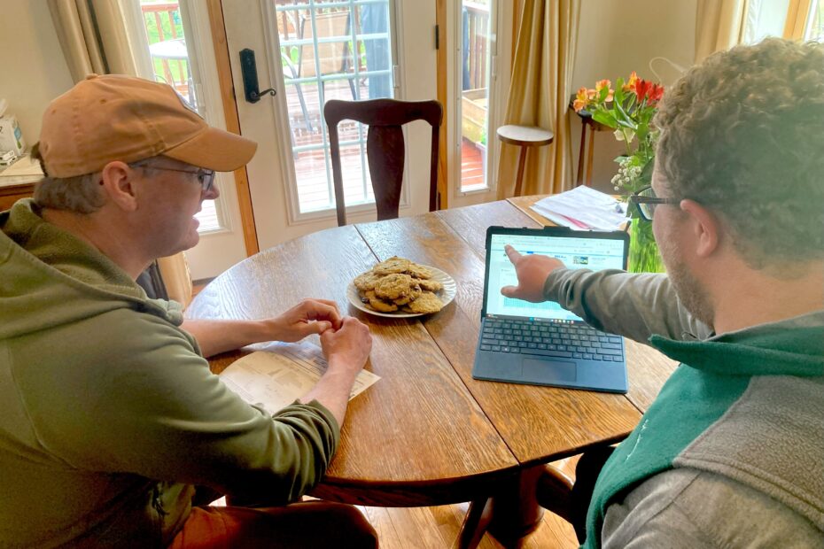 Two people sitting at a table together look at a computer screen. There are fresh-baked cookies on the table.