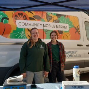 Two people smile in front of the community mobile market van.