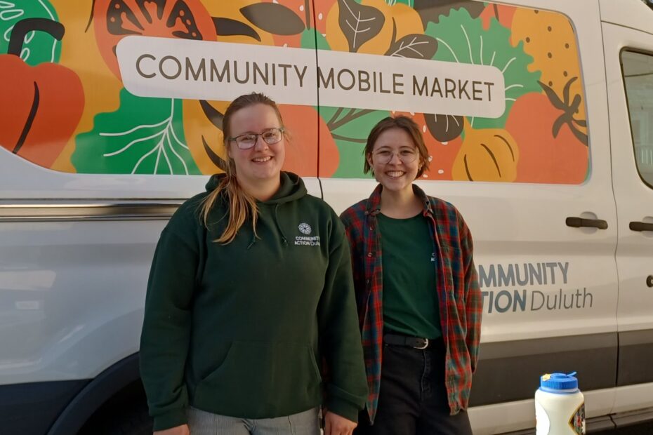 Two people smile in front of the community mobile market van.