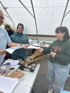 People gather around a table. One person passes garlic bulbs to another person approaching the table. There are garlic bulbs, papers, and planting tools laid out on the table.