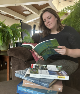 Jenn reading a book, sitting on a couch. In front of her is a pile of other books. She looks very studious indeed.