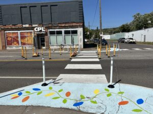 Image of crosswalk installed on Superior St and 23rd Ave. There is a crisp white crosswalk, colorful street mural, and delineators. We can also see delineators and signs make an island in the turn lane, so people crossing have a safe space to stand in the street.