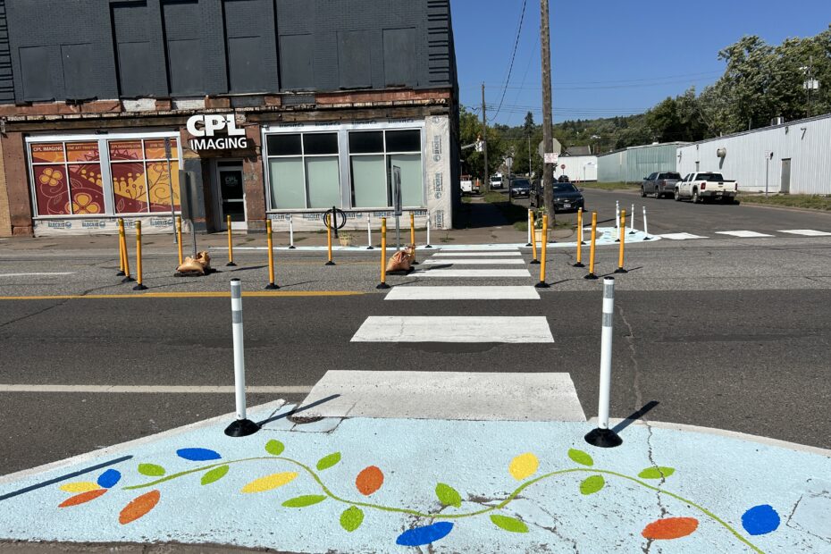 Image of crosswalk installed on Superior St and 23rd Ave. There is a crisp white crosswalk, colorful street mural, and delineators. We can also see delineators and signs make an island in the turn lane, so people crossing have a safe space to stand in the street.