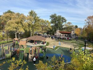 View of Lincoln Park from the hill above. We can see the new playground, full of kids and parents; people sitting under a shade structure, and a large crowd gathered in front of the stone pavilion.