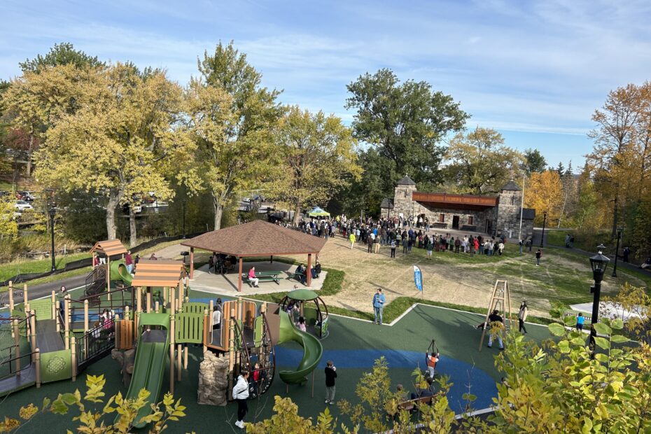 View of Lincoln Park from the hill above. We can see the new playground, full of kids and parents; people sitting under a shade structure, and a large crowd gathered in front of the stone pavilion.