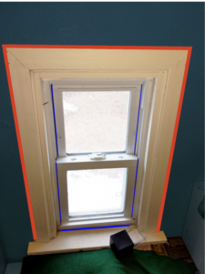 View of a window from inside a house. Blue lines are drawn where the window meets the internal frame. A red line is drawn around the outside of the window trim.