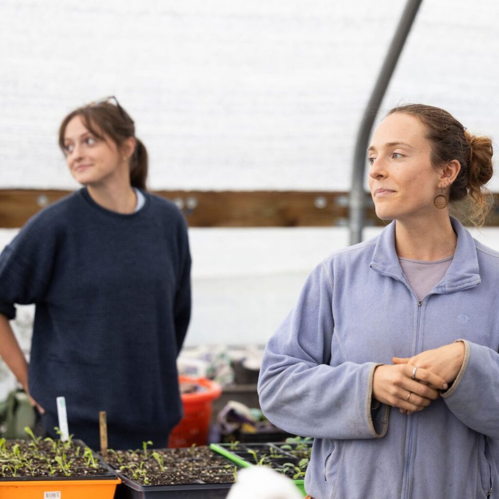 Two people standing in a greenhouse amid seedlings, smiling and looking to the left of the photographer.