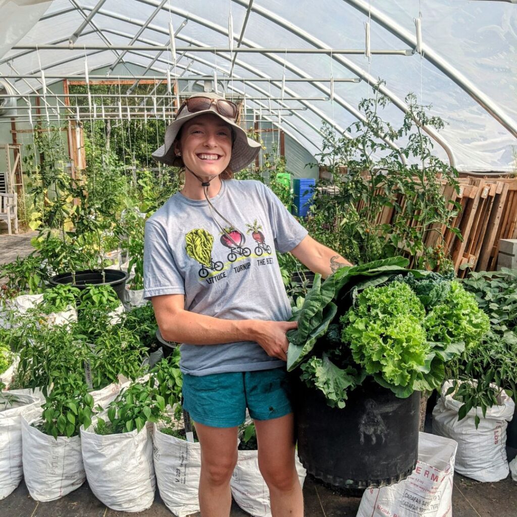 Person smiling in greenhouse, holding a big bucket of gorgeous greens.