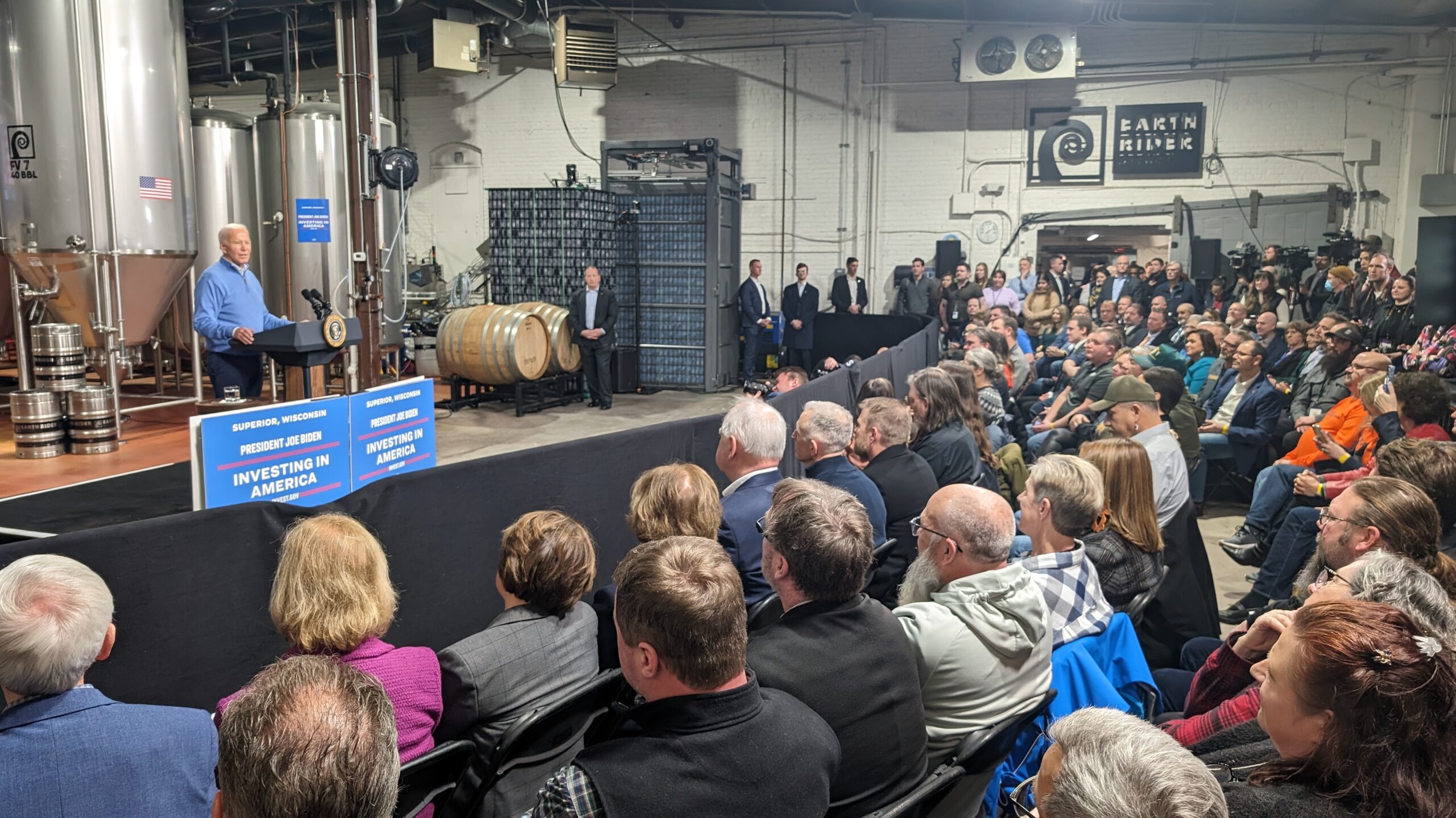 President Biden stands at a podium and speaks to a seated crowd. Brewing equipment takes up the wall behind him.