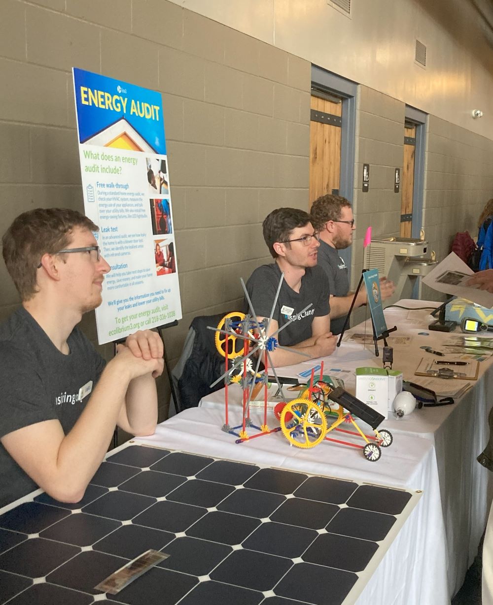 Three people sit behind a table covered in models, papers, and resources about energy efficiency. They are looking off-camera.