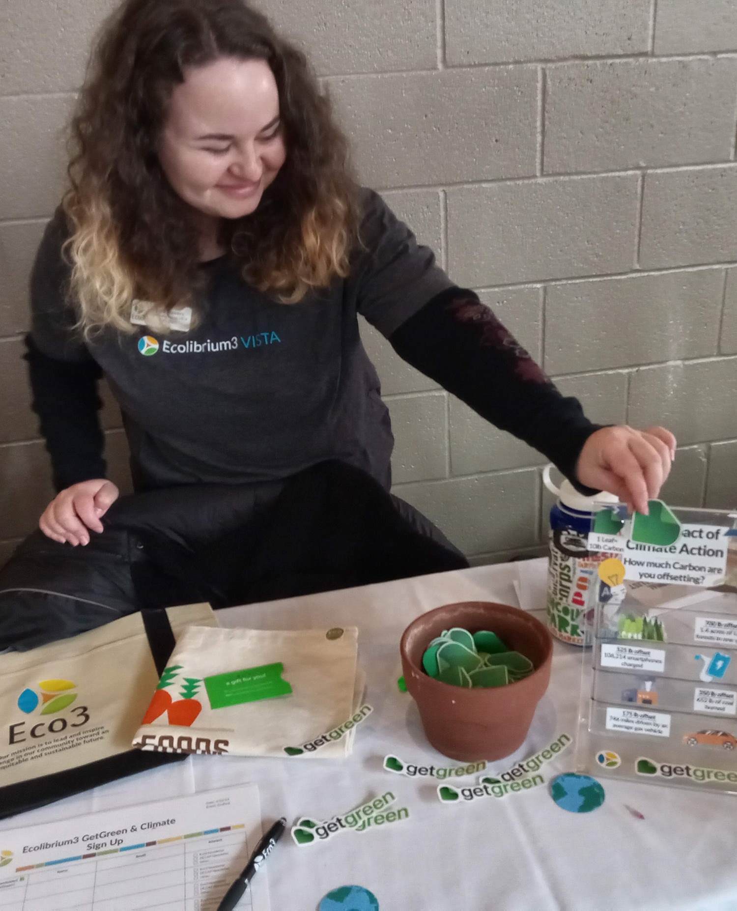 Person puts a small green token into a box, symbolizing climate actions taken. There are stickers, papers, and tote bags laid out on the table.