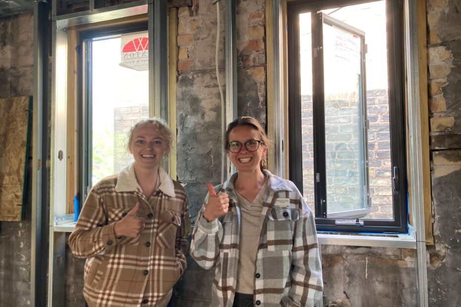 Two people smile and gesture "thumbs up" inside brick building, in front of two windows.