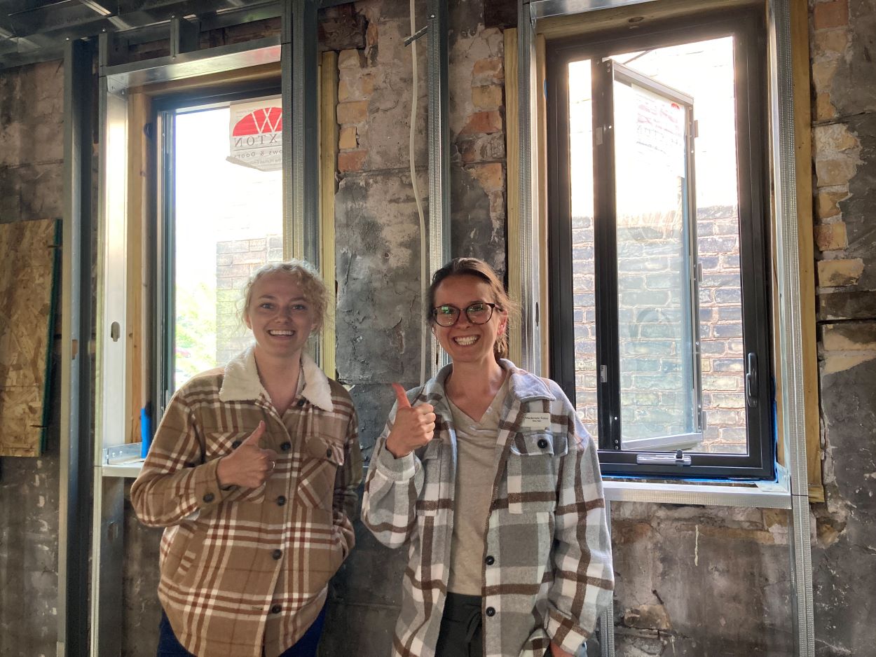 Two people smile and gesture "thumbs up" inside brick building, in front of two windows.
