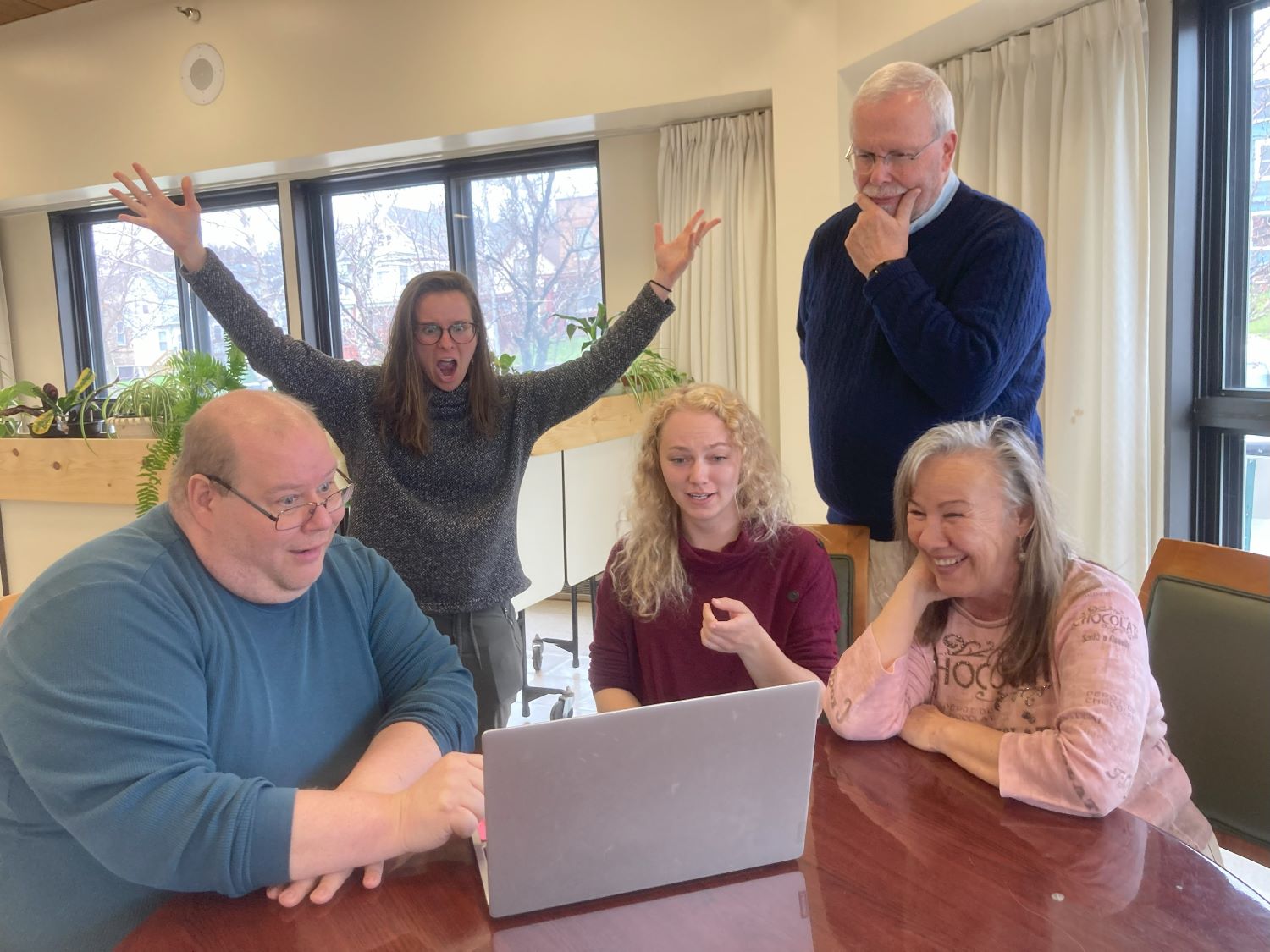 Five people gathered around a laptop show joy and surprise in this silly photo.