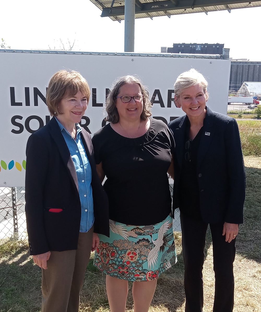 Three people pose for photos in front of the Lincoln Park Solar Garden sign.