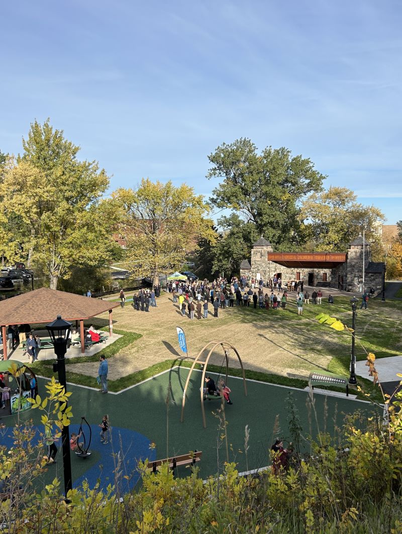 Overview of park on sunny day, crowd of people gathered in front of a pavilion.