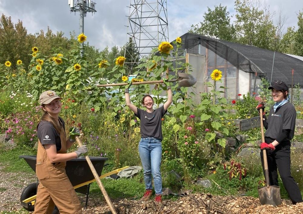 Three smiling people pose with shovels and rakes in front of a greenhouse and a flower bed on a sunny day.