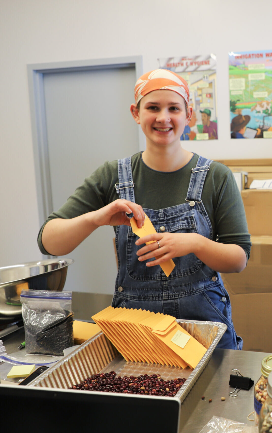 Person wearing overalls smiles as they put small round seeds into envelopes.