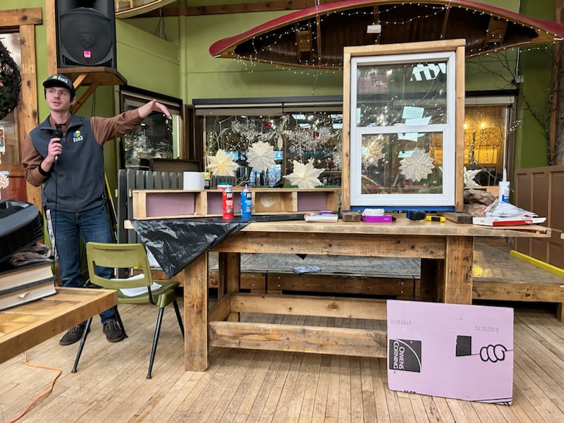 A person wearing an Eco3 vest and hat points at a window and other demonstration tools on a table. He is speaking into a microphone.