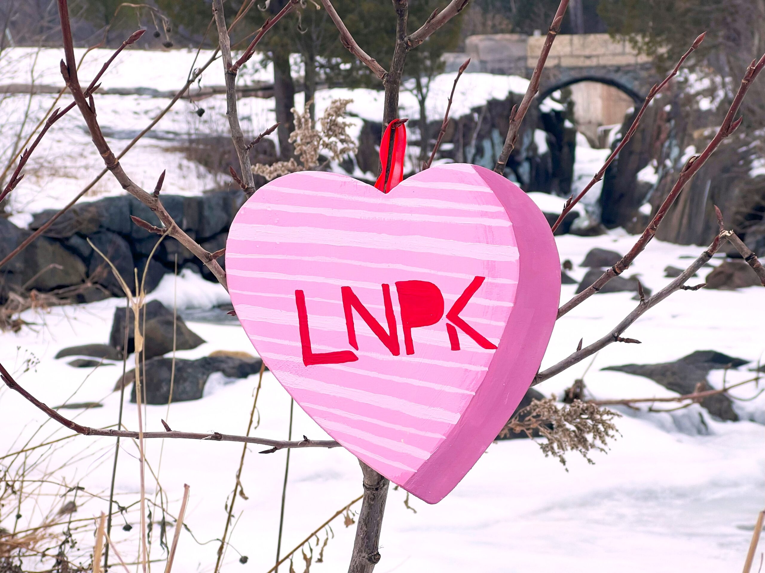 Pink heart with stripes and "LNPK" in red hangs on a tree in front of a frozen creek.