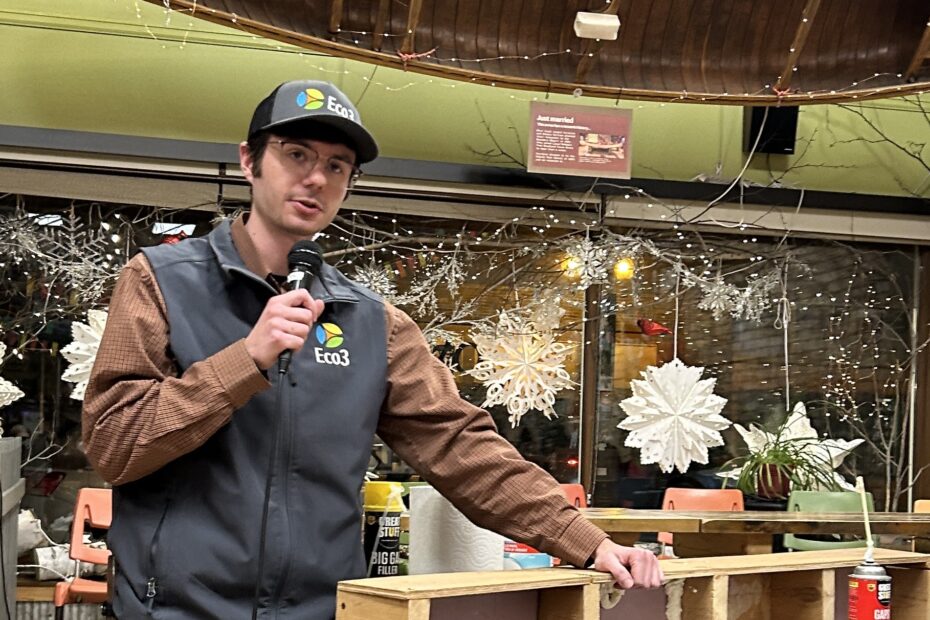 Person with Eco3 hat and vest speaks into a microphone while leaning on a table with demonstration materials on it.