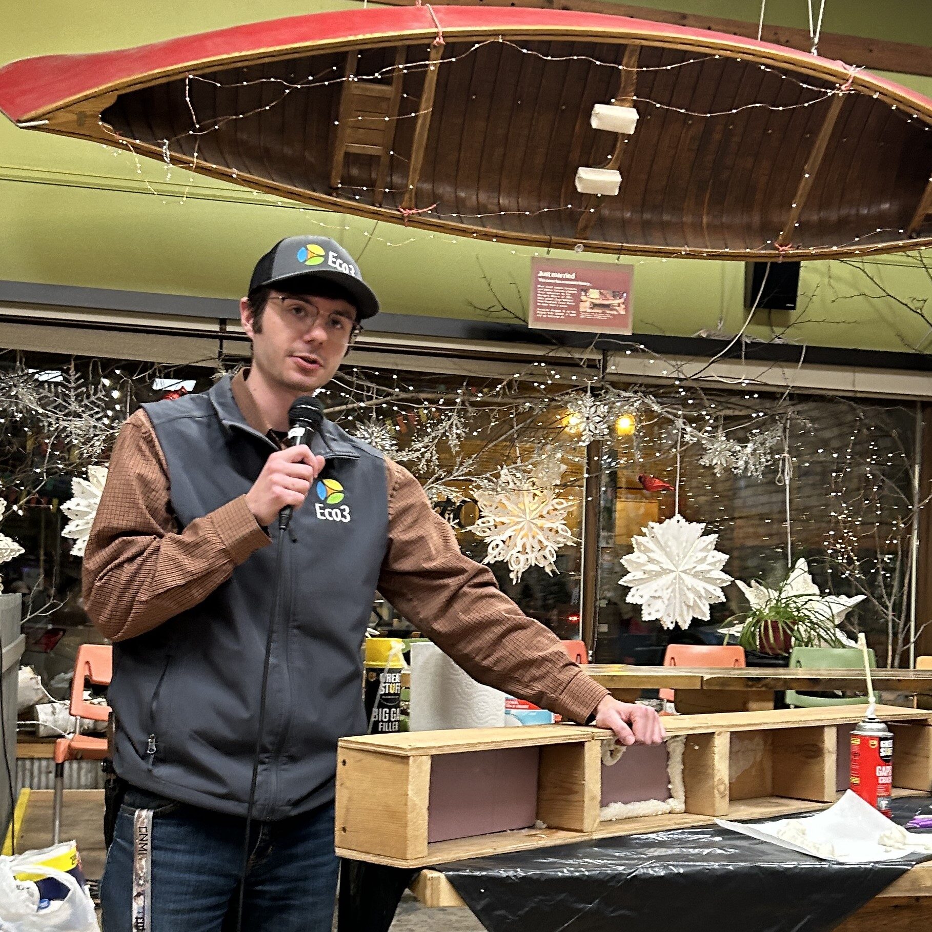 Person with Eco3 hat and vest speaks into a microphone while leaning on a table with demonstration materials on it.