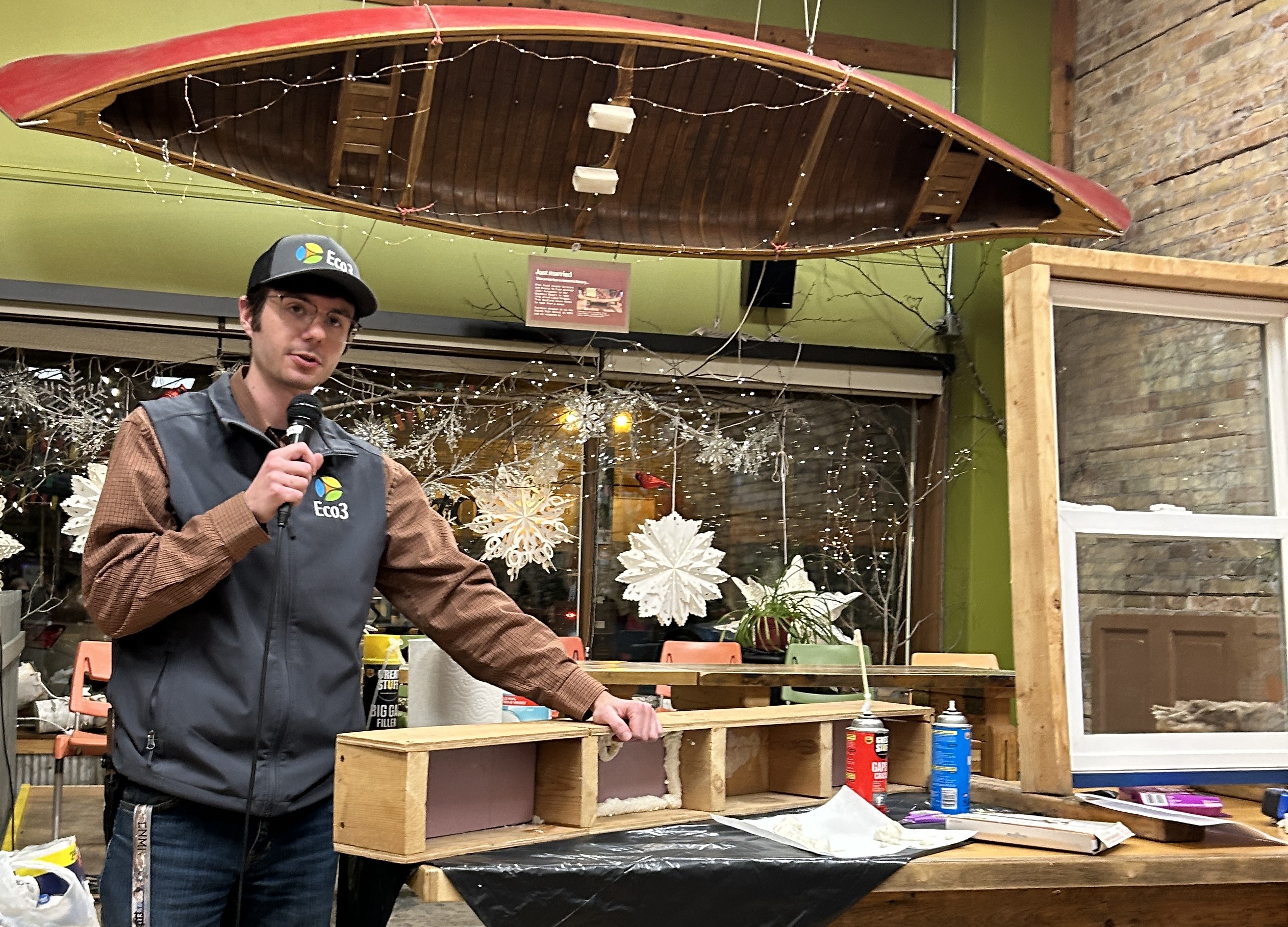 Person with Eco3 hat and vest speaks into a microphone while leaning on a table with demonstration materials on it.