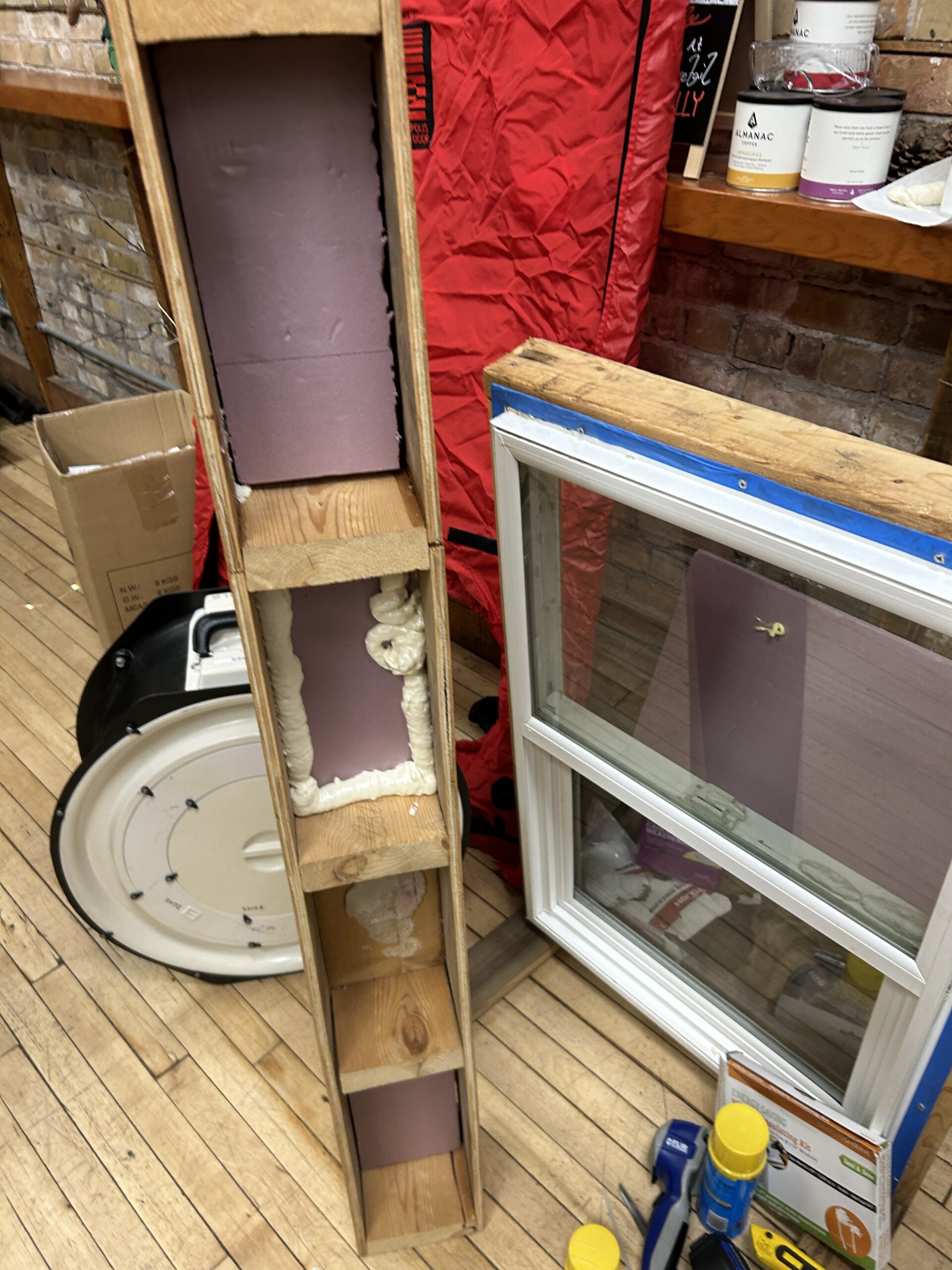 Tools and demonstration materials grouped on the floor: wood with spray foam insulation, a stand-alone window, an infrared camera, and a box of window kits.