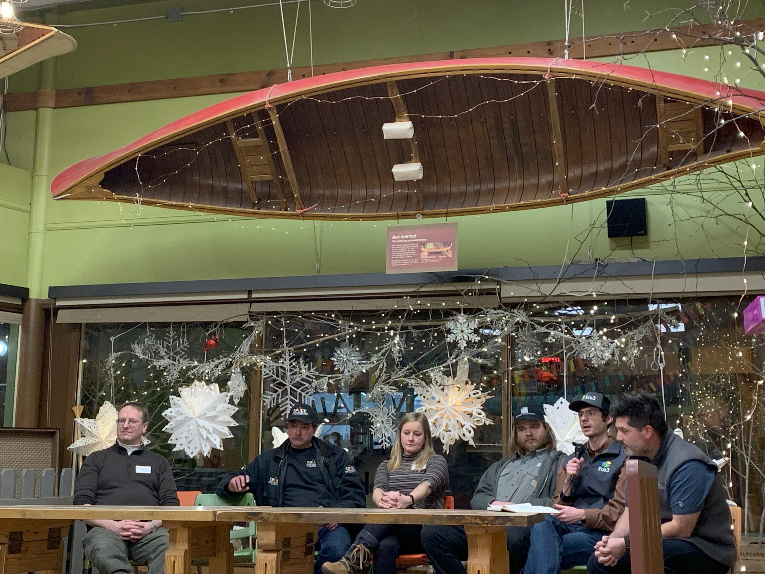 Six people sit around a table, facing the camera. One person is speaking into a microphone.