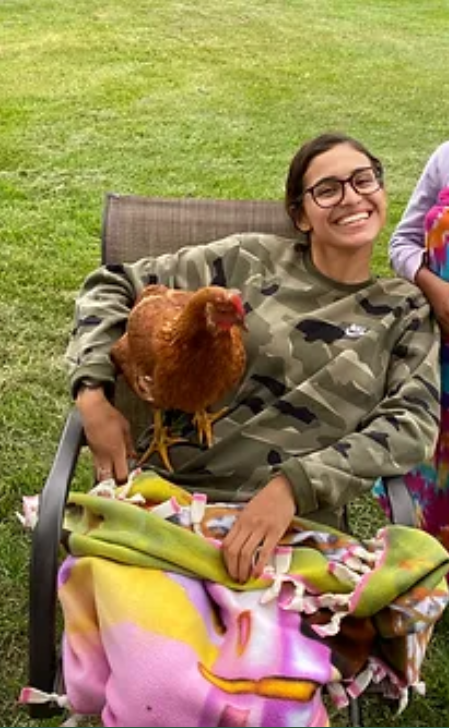 A person sits in a lawn chair, smiling at the camera, with a chicken on her lap.