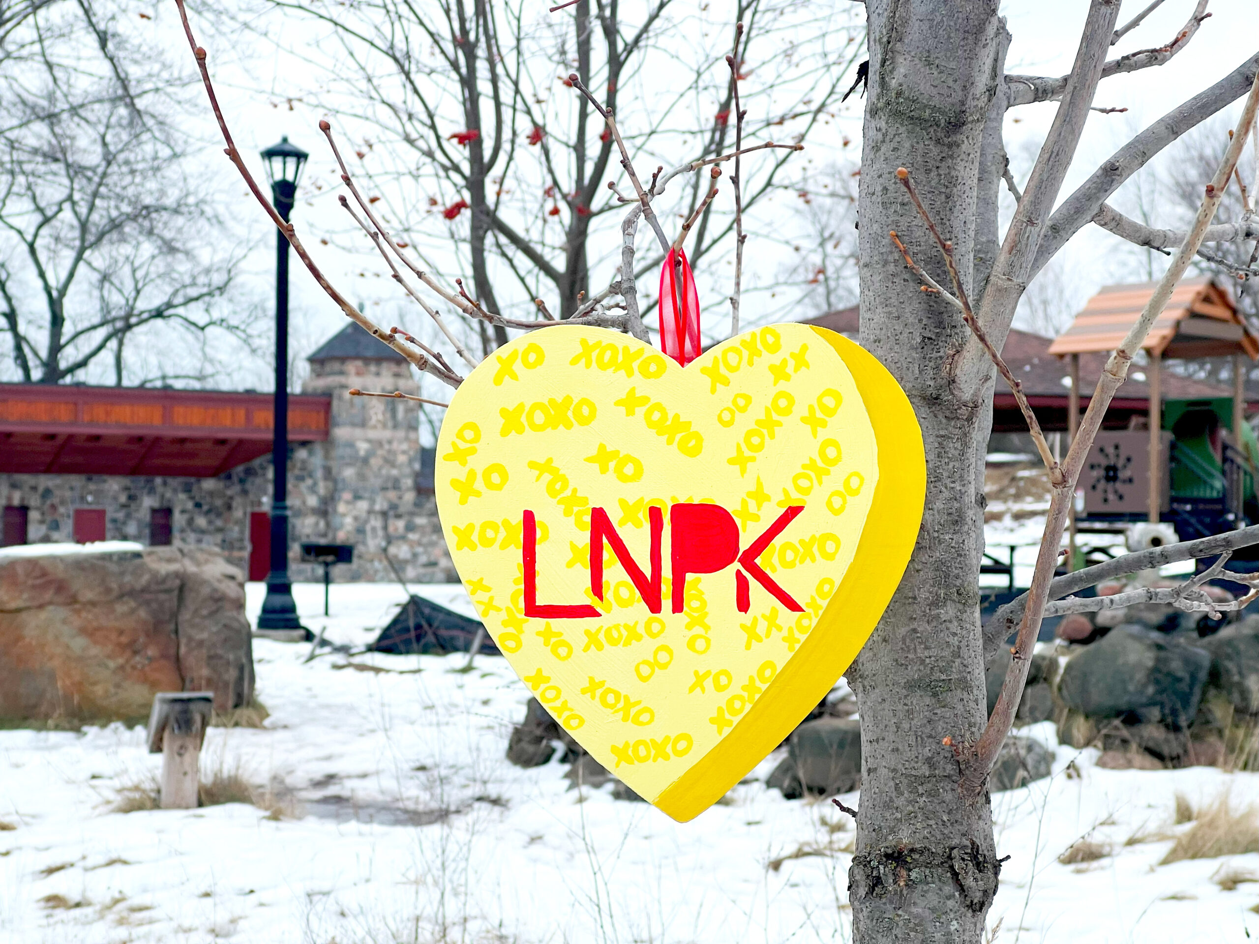 Yellow heart with XOXO pattern and "LNPK" in red hangs on a tree in front of the Lincoln Park pavilion.