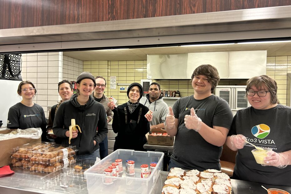 Seven smiling people stand behind the counter in a commercial kitchen. Some are wearing t-shirts that say "Eco3 VISTA." On the counter there are breakfast foods laid out.