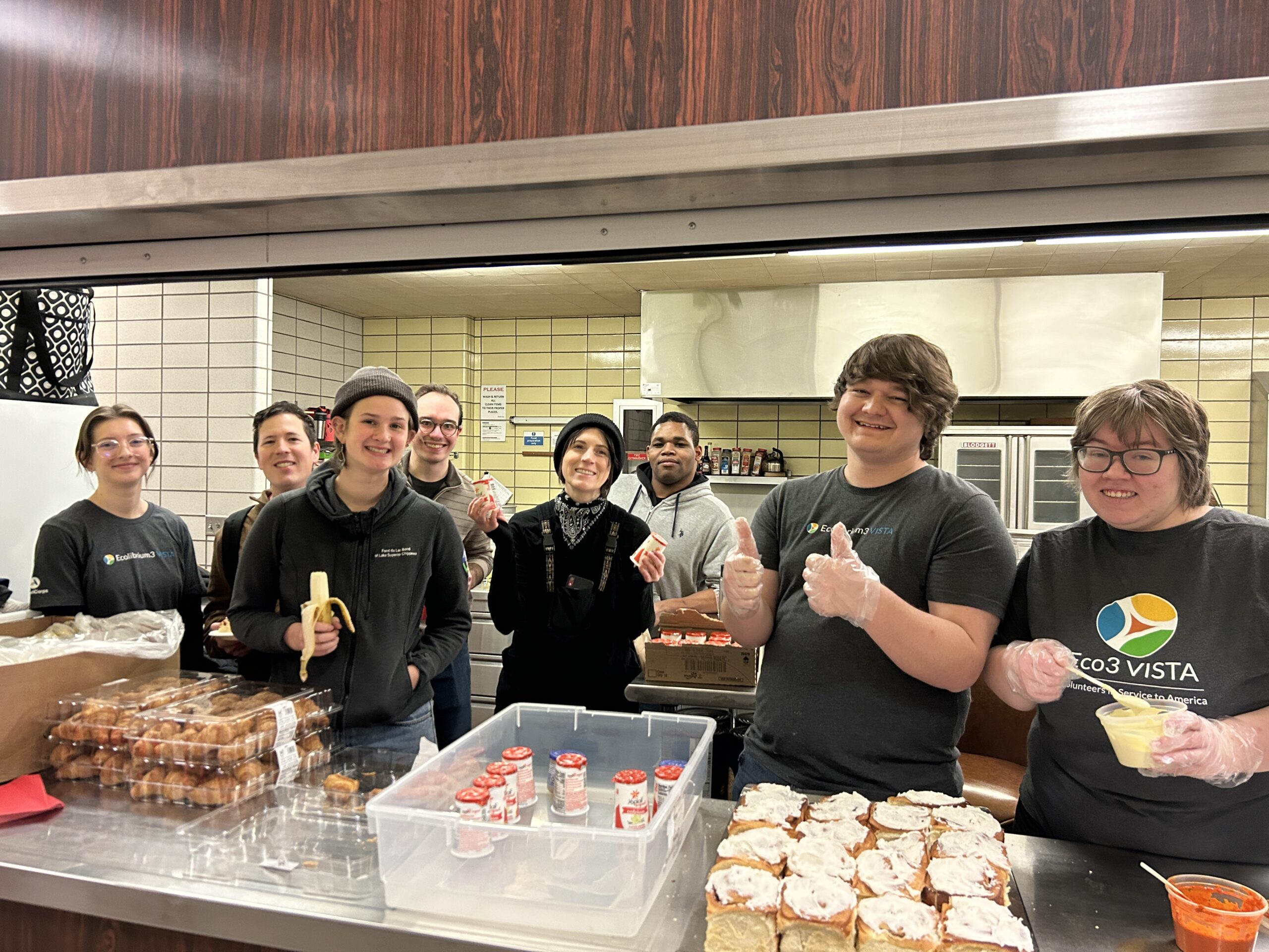 Seven smiling people stand behind the counter in a commercial kitchen. Some are wearing t-shirts that say "Eco3 VISTA." On the counter there are breakfast foods laid out.