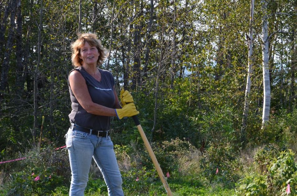 Person with shovel smiles in a field in front of trees.